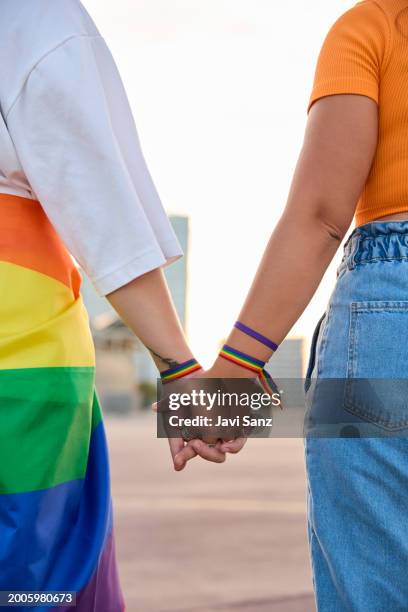 foto vertical de una pareja de lesbianas irreconocibles tomados de la mano con un brazalete arcoíris celebrando el día del orgullo lgbt. - desfile del orgullo gay fotografías e imágenes de stock