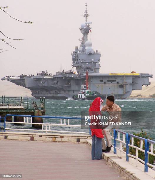 An Egyptian couple stands on a pier as the French Nuclear aircraft carrier Charles de Gaulle navigates in the Suez Canal in front of the port city of...