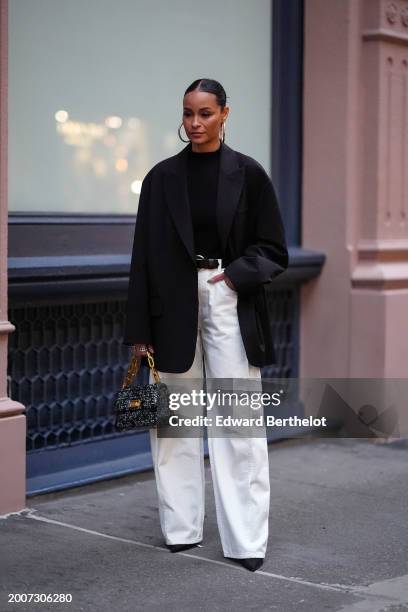 Sai de Silva wears earrings, a black oversized blazer jacket, a black top, a belt, white flared pants , pointed shoes , a bag, outside Tibi, on...