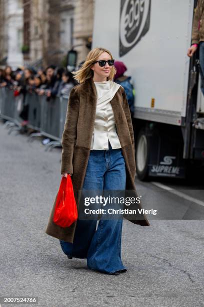 Lisa Aiken wears wide leg denim jeans, brown coat, red bag, silk top outside Coach on February 12, 2024 in New York City.