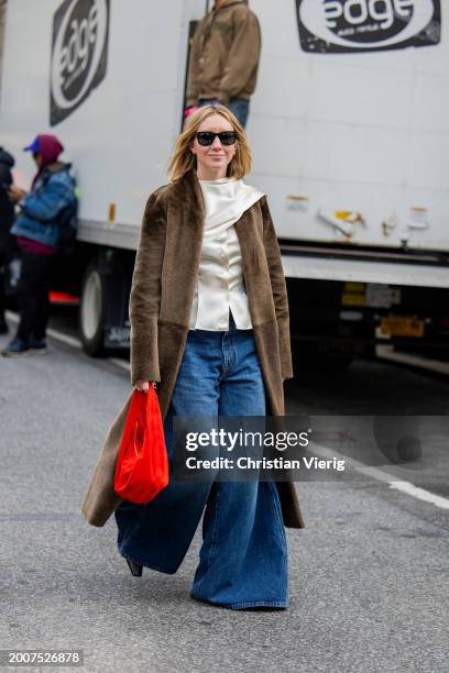 Lisa Aiken wears wide leg denim jeans, brown coat, red bag, silk top outside Coach on February 12, 2024 in New York City.
