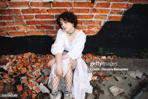 sad young woman sitting in ruins by the wall among broken bricks - shelling stock pictures, royalty-free photos & images