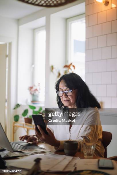 senior woman using mobile phone and laptop while sitting at home - occhiali da lettura foto e immagini stock