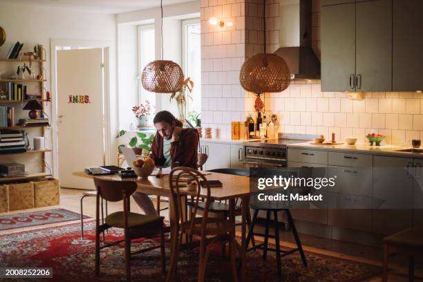 young man using laptop while sitting at dining table in kitchen - study table stock pictures, royalty-free photos & images
