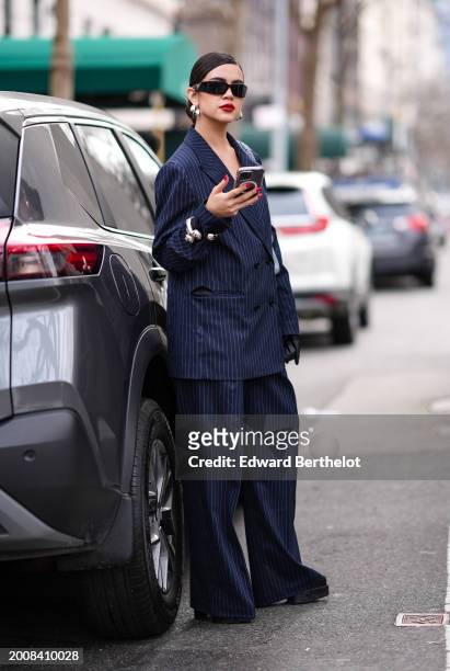 Guest wears sunglasses, silver earrings, a blue striped oversized blazer jacket , matching flared pants , red lipstick, make-up , holds a mobile...