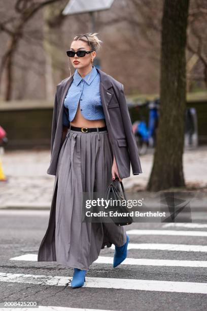 Guest wears earrings, sunglasses, a blue cropped shirt, a gray oversized blazer jacket, a Coach belt, a long slit pleated gray skirt, a black leather...