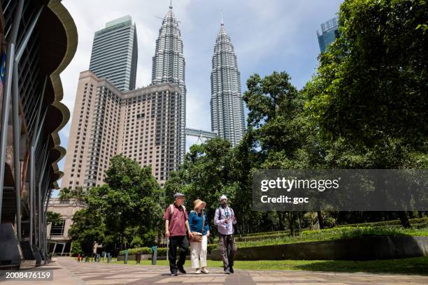 full length view of a mature sikh tour guide leading two senior asian tourist through the famous kuala lumpur petronas twin towers landmark in the kuala lumpur city centre, malaysia. - indian tour guide stock pictures, royalty-free photos & images