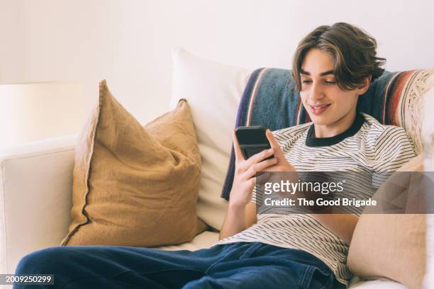 smiling teenage boy using smart phone while sitting on sofa at home - malibu home stock pictures, royalty-free photos & images