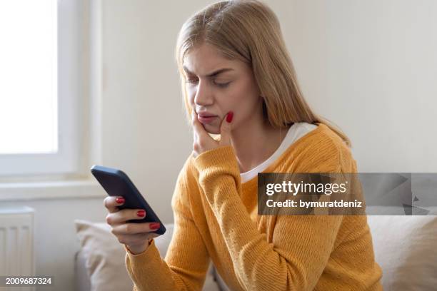 worried woman sitting on sofa at home and holding phone - scam victim stock pictures, royalty-free photos & images