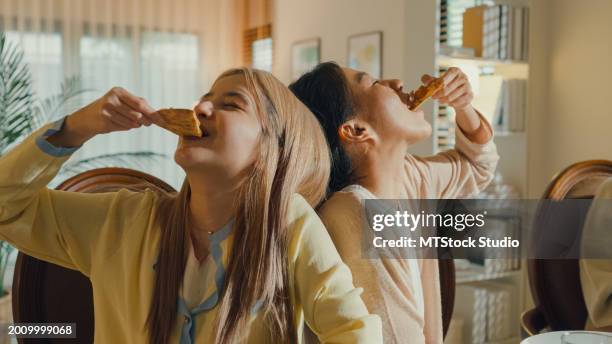 group of young asian people eating pizza and having fun sitting at dining table at home. multicultural friends enjoying spending together college house party. - biting stock pictures, royalty-free photos & images