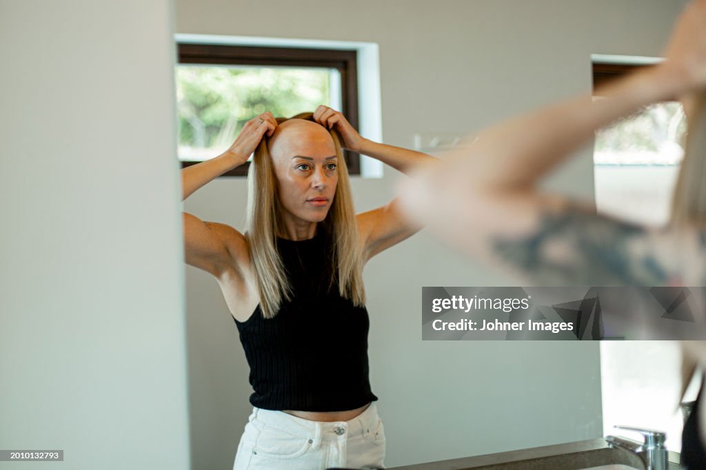 Reflection of bald woman wearing wig standing in bathroom at home