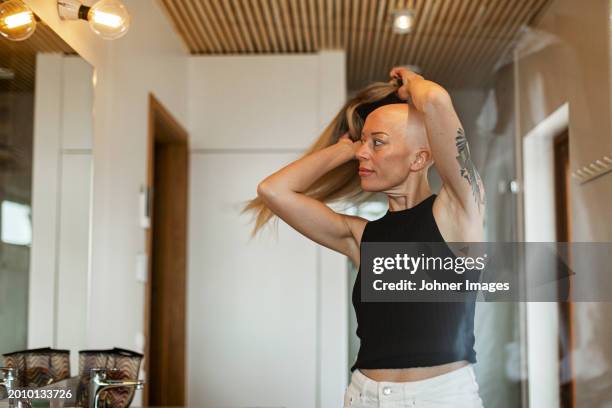 bald woman suffering from cancer wearing wig while looking in mirror at home - wig stock pictures, royalty-free photos & images