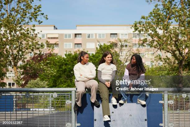 multiracial female friends talking while sitting on railing at sports court during sunny day - school railings stock pictures, royalty-free photos & images