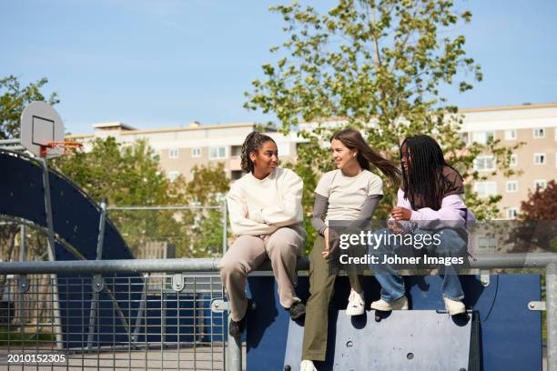 teenage girl talking with female friends while sitting on railing during sunny day - school railings stock pictures, royalty-free photos & images