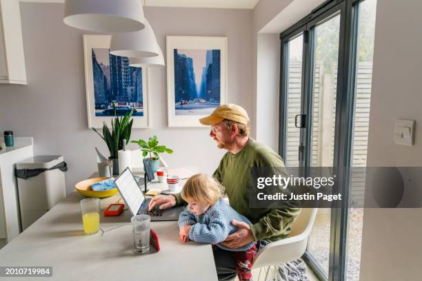 side view of a mature red headed man working from home with his baby daughter on his lap - flexible working stock pictures, royalty-free photos & images