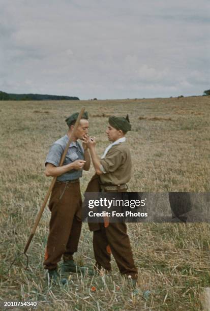 German prisoners of war, one holding a pitchfork as the other lights a cigarette for him as the work on farmland in England, United Kingdom, circa...