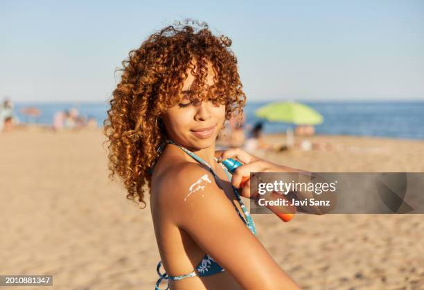 cheerful young woman on the beach applies sunscreen on her shoulder - bronzeado imagens e fotografias de stock
