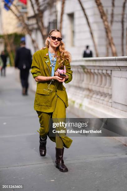 Guest wears sunglasses, earphones, a blue pullover, a green / khaki oversized blazer jacket, a khaki lustrous shiny silk slit skirt , brown leather...