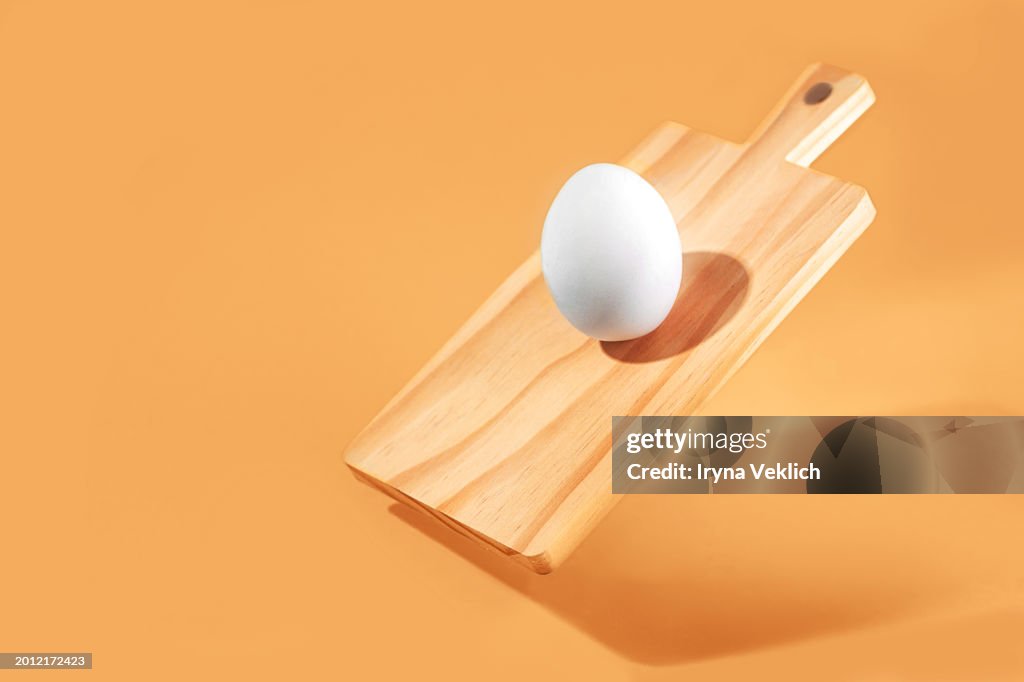 Creative Easter design concept with White Easter egg on wooden kitchen cutting board on pastel color trend 2024 peach color table background.