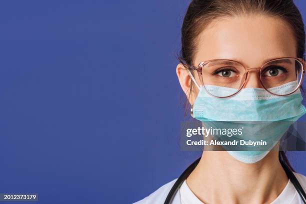 closeup portrait of a woman doctor wearing a medical face mask on a blue background studio shot - studio head shot serious confident looking at camera imagens e fotografias de stock