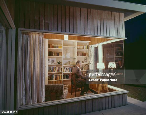 View through a large glass window of a man working at a desk in a home office cum library in a modern house in the village of Stapleford in...