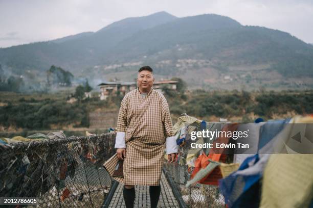 portrait bhutanese mature man looking at camera standing on bridge in front of village - thimphu 個照片及圖片檔