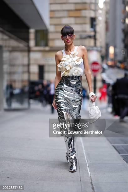 Katya Tolstova wears sunglasses, golden earrings , red lipstick , a white tank top with floral attachments, a silver shiny midi skirt, high matching...