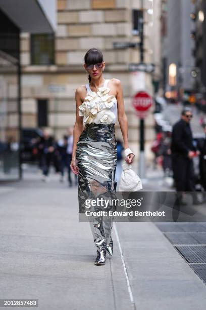 Katya Tolstova wears sunglasses, golden earrings , red lipstick , a white tank top with floral attachments, a silver shiny midi skirt, high matching...