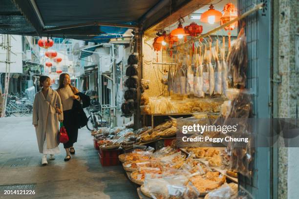 two asian young women are walking past a store lined with a variety of packaged food items, dried goods such as seafood, meats, and snacks, displayed on tables and hanging from the storefront in hong kong - fish market stock pictures, royalty-free photos & images