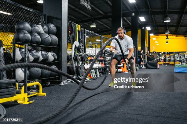young man training with battle rope while listening to music in the gym - cross training foto e immagini stock