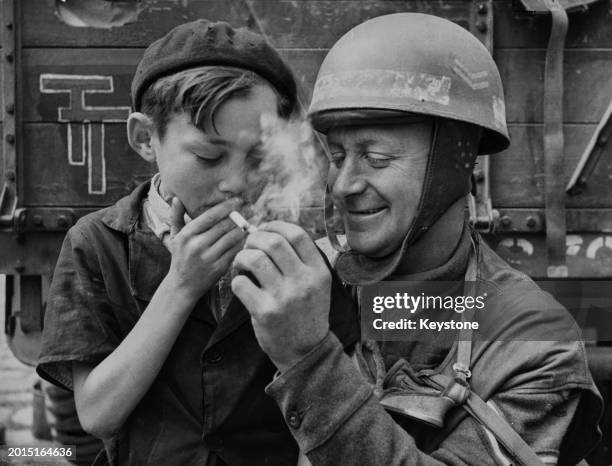 Corporal Fred Sicklong, a motorcycle despatch rider from XXX Corps, British 2nd Army lights a cigarette for a young French boy in the liberated town...