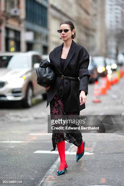 Guest wears earrings, sunglasses, a black oversized blazer jacket, a belt, a black leather puff large bag, red leggings, pointed shoes, outside Tibi,...