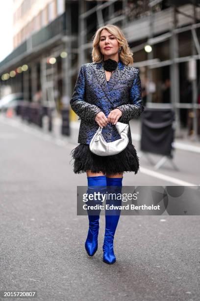 Olga Ferrara wears a black choker with floral detail, a blue shiny double breasted oversized blazer jacket with printed patterns, a black fluffy mini...