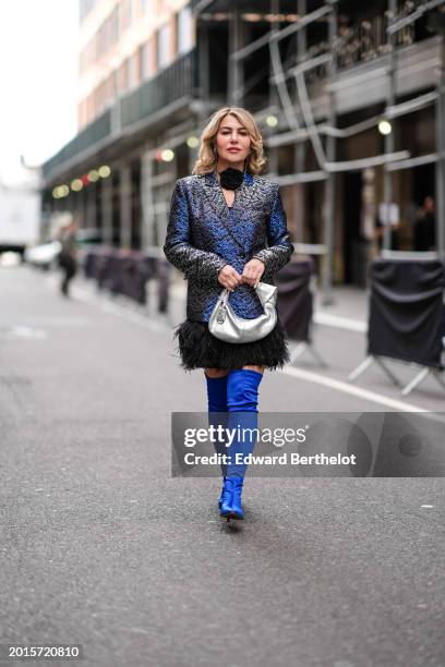 Olga Ferrara wears a black choker with floral detail, a blue shiny double breasted oversized blazer jacket with printed patterns, a black fluffy mini...