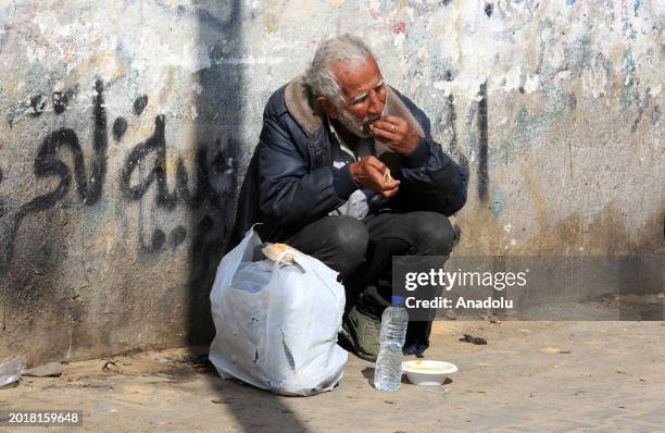 Palestinian man sits in front of wall and eats the distributed food he barely receive, as volunteers prepare hot meal for Palestinian families...