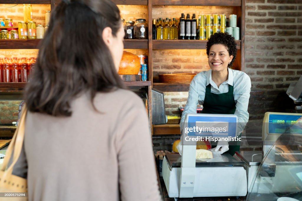 Cheerful saleswoman weighting a cheese on scale for an unrecognizable female customer at the deli
