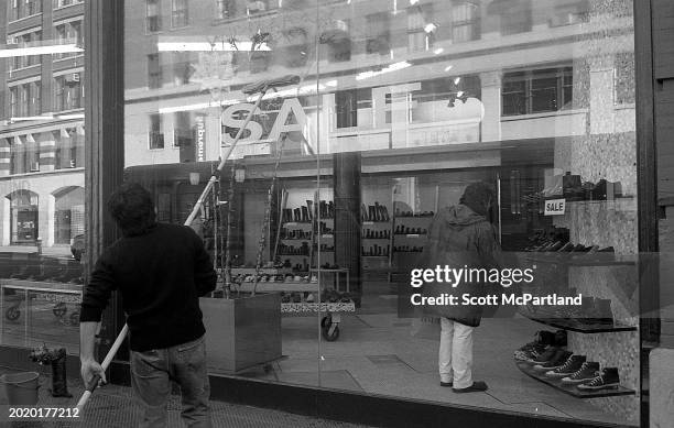 Man cleans the exterior windows of a shoe store while, inside, another looks a boots, on 5th Avenue in midtown Manhattan, New York, New York,...