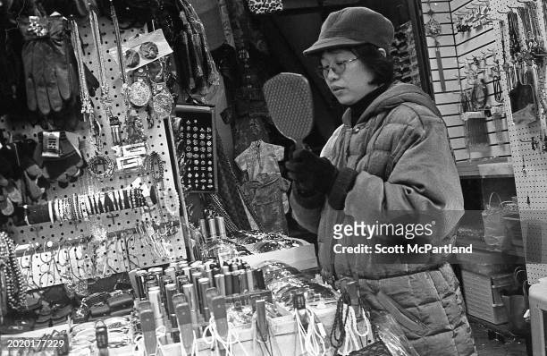 Woman uses a hand mirror to check her reflection as she tries on a pair of glasses at a sidewalk vendor's stall on Canal Street, in the Chinatown...