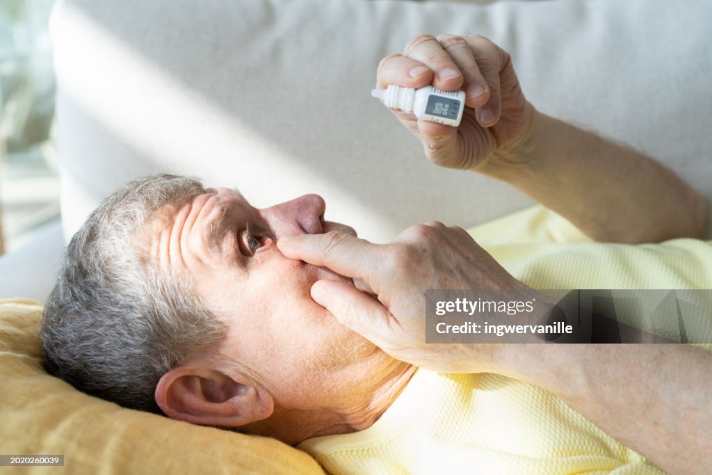 Close-up of senior man putting eye drop while lying on sofa