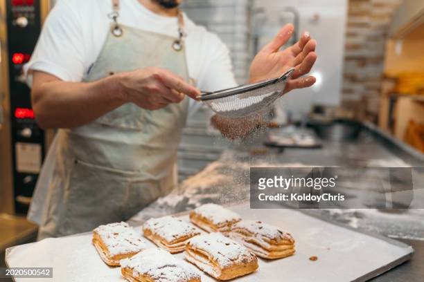 baker dusting powdered sugar on pastry in a bakery kitchen - powdered sugar sifter fotografías e imágenes de stock