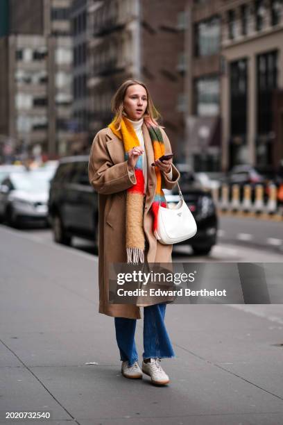 Guest wears a beige long coat, a colored wool fringed striped scarf, a white leather Prada bag, blue denim jeans pants , white sneakers shoes,...