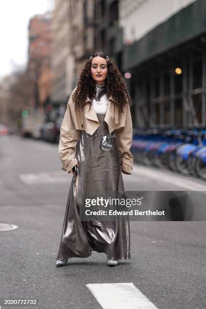 Guest wears hair clips, a beige cropped short trench coat , a turtleneck long shiny silver slit dress , matching boots , a matching crossbody bag,...