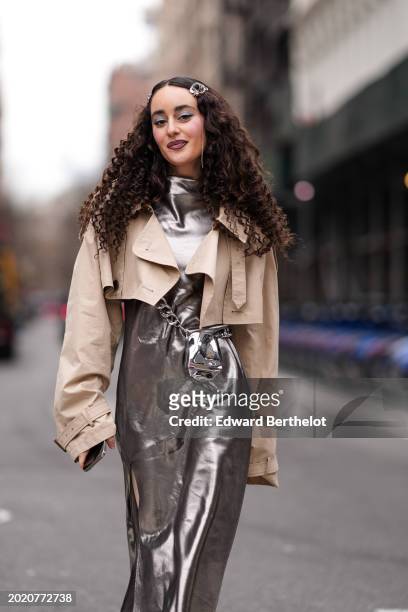 Guest wears hair clips, a beige cropped short trench coat , a turtleneck long shiny silver slit dress , matching boots , a matching crossbody bag,...