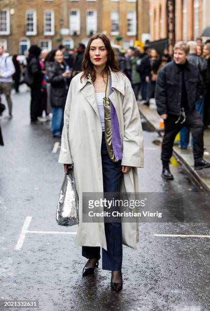 Alexa Chung wears trench coat, silver sequined bag, blue striped pants outside JW Anderson during London Fashion Week February 2024 on February 18,...