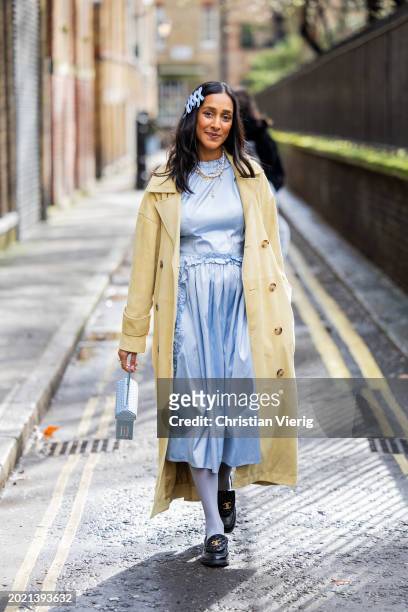 Zeena Shah wears blue bows in hair, blue dress, pearl necklace, beige coat, Chanel loafers, tights, blue bag outside Eudon Choi during London Fashion...