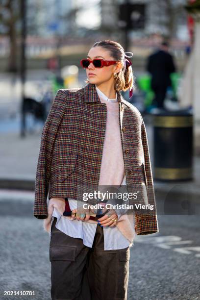 Gabby Martin wears red Prada sunglasses, checkered jacket, oversized knit, grey pants with side pockets outside Holzweiler during London Fashion Week...