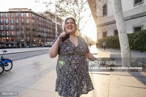curvy woman happy in the city listening to music headphones smart phone singing - voluptuoso fotografías e imágenes de stock
