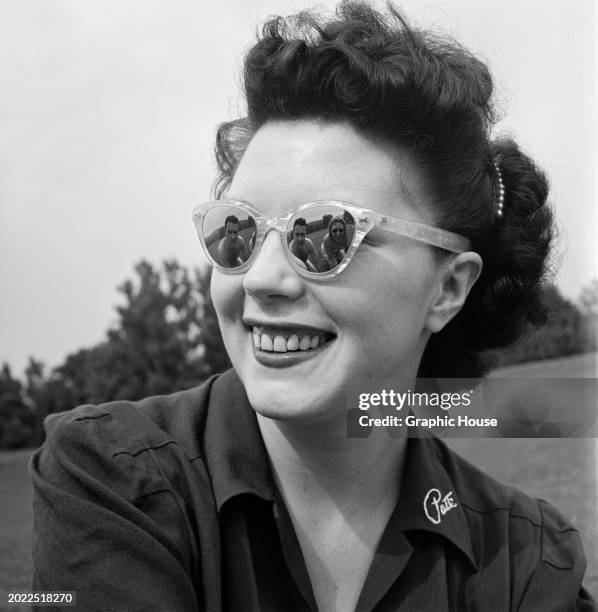 Smiling model wearing sunglasses - with people reflected in the lenses - and a dark shirt with an open collar, United States, circa 1950.