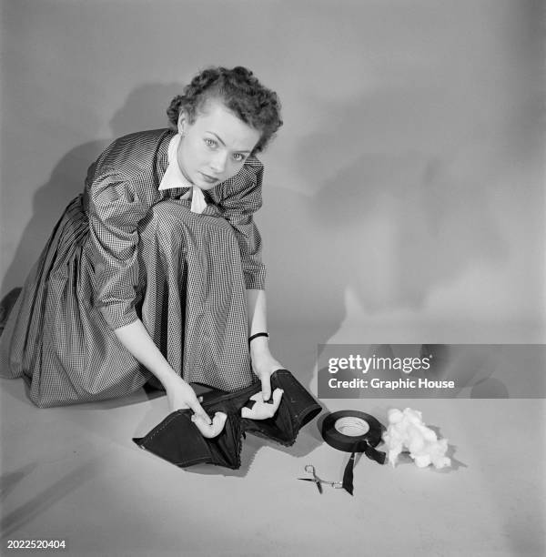 Model crouches to prepare a strapless bra, holding the bra in her hands, with cotton wool, tape, and a pair of scissors on the ground before her,...