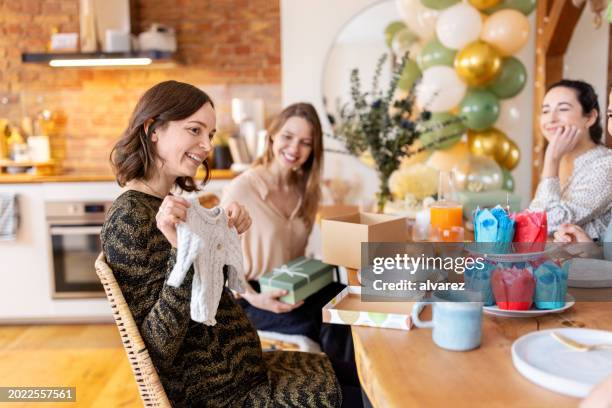 pregnant woman opening presents and showing them to friends sitting by during her baby shower - chá de bebé imagens e fotografias de stock